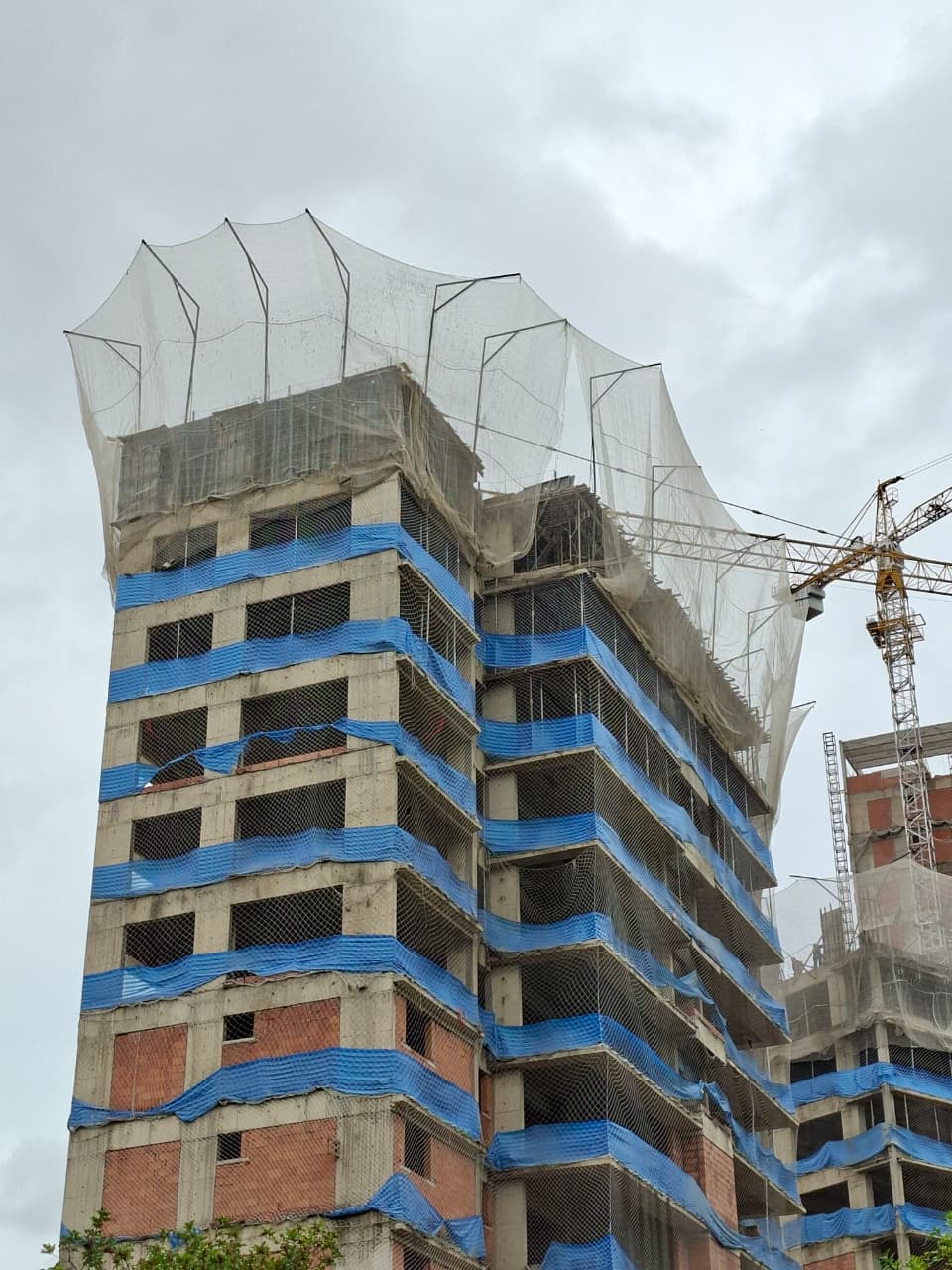 Prédio em construção com rede de proteção SLQA no topo e telas azuis nos pavimentos, guindaste ao lado