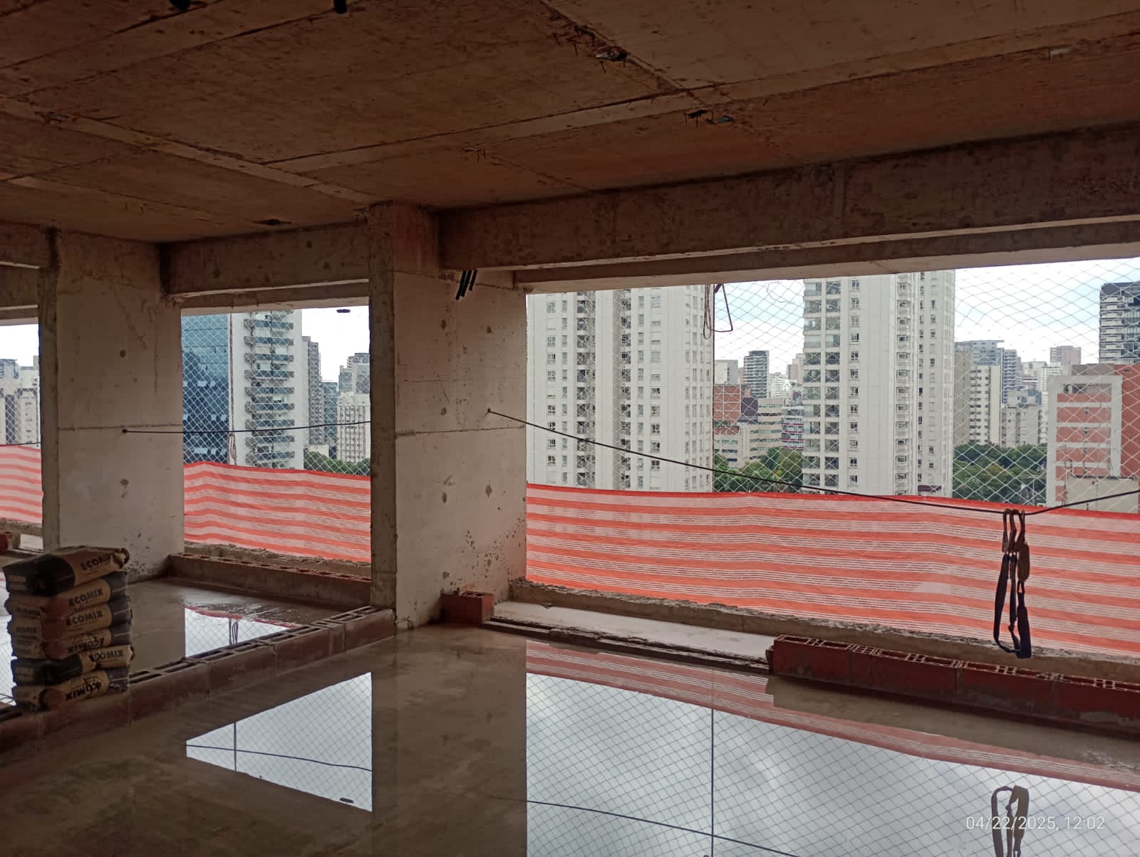 Pavimento de obra em construção com tela alaranjada de proteção na abertura e vista para prédios da cidade